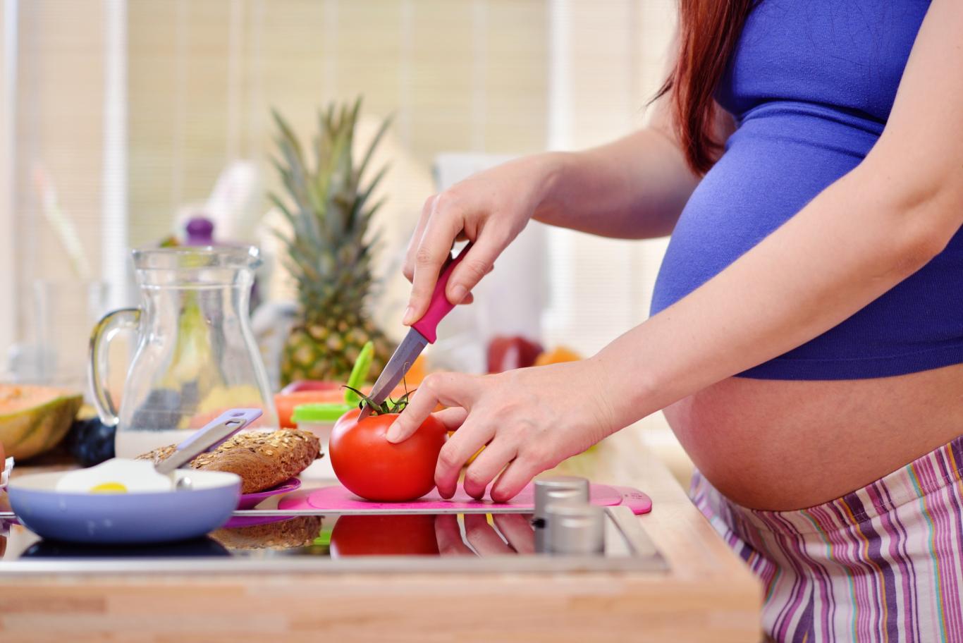 Las manos de una mujer embarazada cortan un tomate sobre la encimera de la cocina
